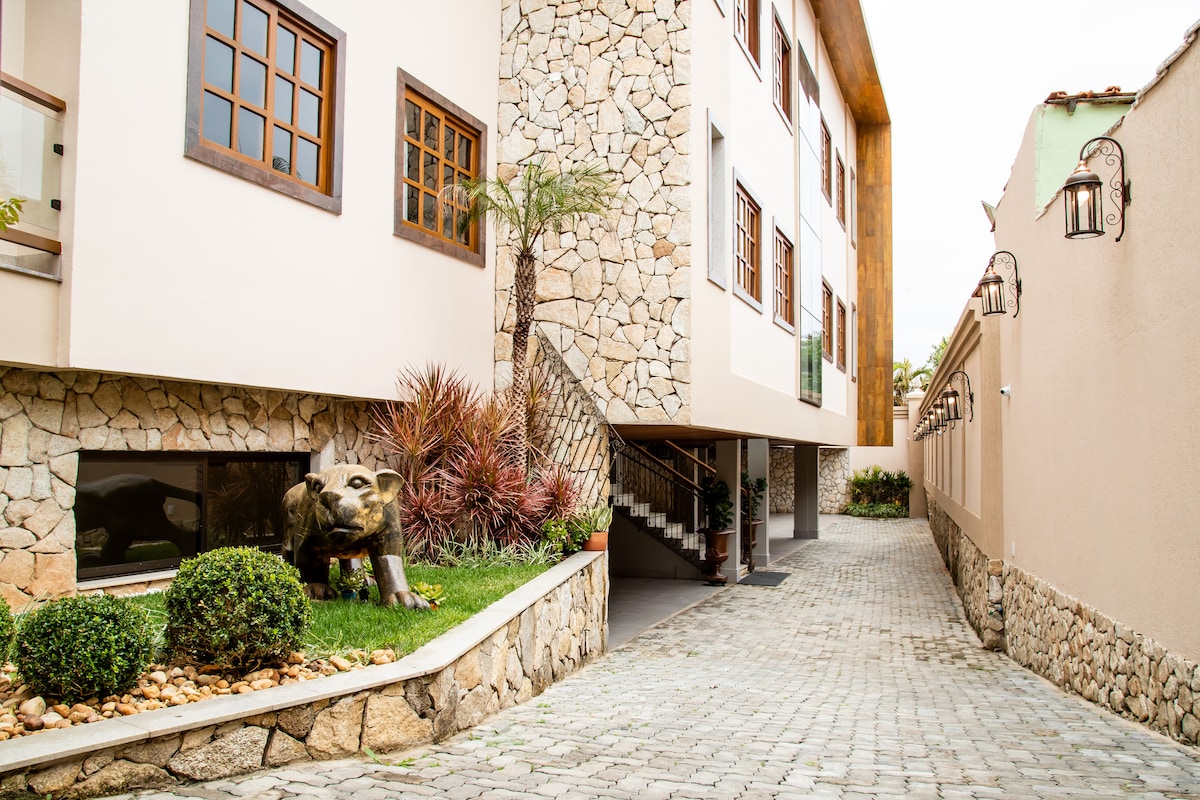 A pathway leads to the entrance of a modern building, flanked by well-maintained landscaping. Decorative stone walls and a lion sculpture are visible, adding character and charm to the exterior. The entrance is enhanced by subdued architectural details, contributing to a welcoming ambiance.