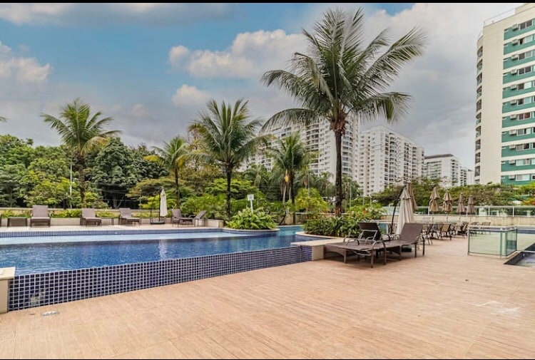 A well-maintained pool area features a sparkling blue swimming pool surrounded by palm trees. Comfortable lounge chairs are arranged along the poolside, with umbrellas providing shade. Lush greenery enhances the tranquil atmosphere, complemented by modern apartment buildings in the background.