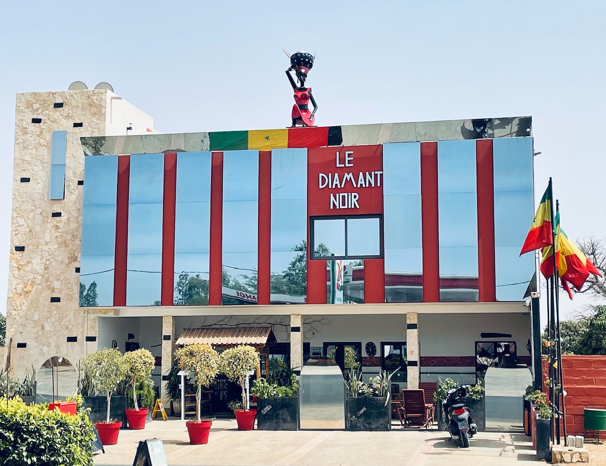 The exterior of Le Diamant Noir showcases a modern design with large glass panels reflecting the surroundings. A colorful Senegalese flag is displayed prominently, alongside two trees in decorative pots, flanking the entrance. A unique sculpture stands atop the building, adding an artistic touch.
