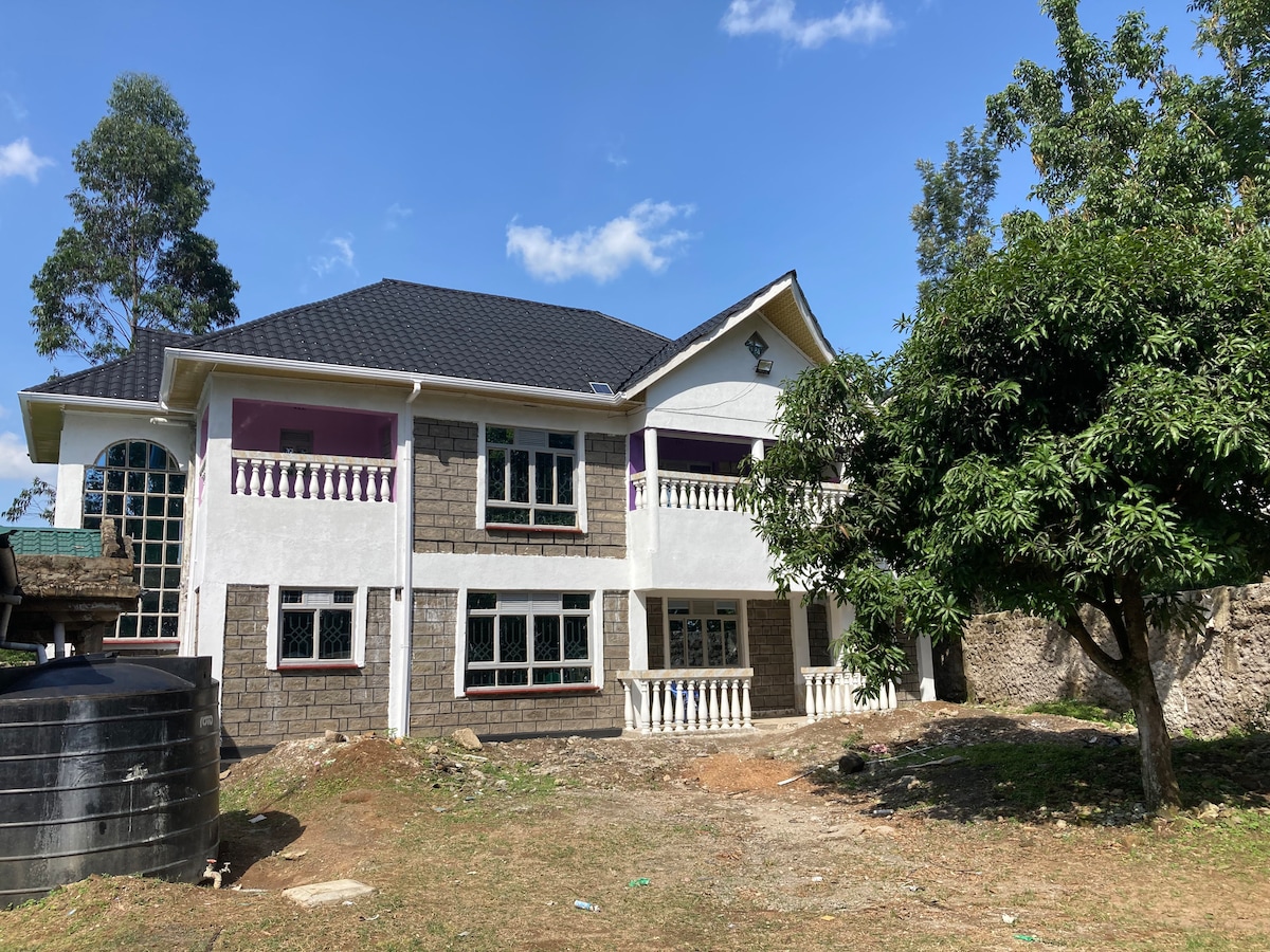 A two-story house with a mix of stone and plastered exterior features large windows and decorative balconies. The lush green landscape surrounds the home, with a tree visible in the foreground and a water tank nearby. Clear blue skies complement the inviting exterior.