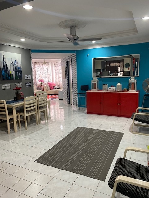 A spacious living area features bright blue walls and white tiled flooring. A dining table with six chairs sits beside a red cabinet. The room is complemented by a striped area rug and a ceiling fan. In the background, another room is partially visible.