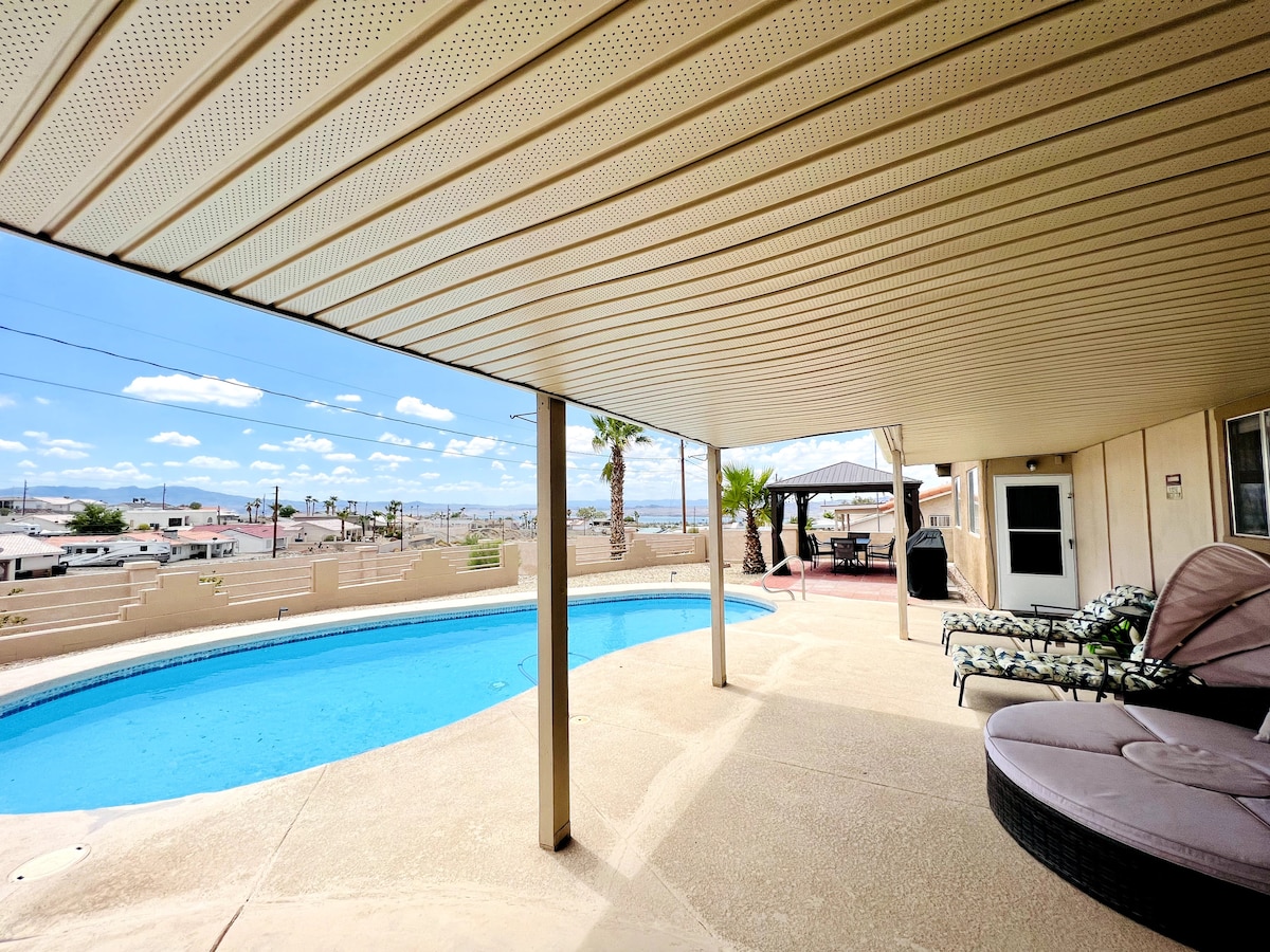 A covered patio features a clear blue pool surrounded by a spacious sun deck. Lounge chairs are positioned nearby, with palm trees and a scenic view in the background. Bright, open skies contribute to the light-filled atmosphere.