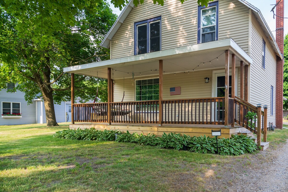 A welcoming porch area features wooden railings and ample seating under soft string lights, providing a relaxed outdoor space. The home, surrounded by lush greenery, includes a well-maintained lawn. A large window allows natural light to brighten the interior.
