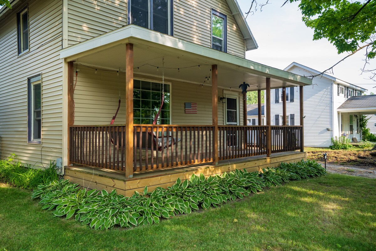 A spacious porch is shown with a wooden railing and swing chairs, surrounded by lush green foliage. The porch is illuminated by string lights overhead, creating a cozy outdoor space. An American flag is displayed on the wall, adding a touch of charm.