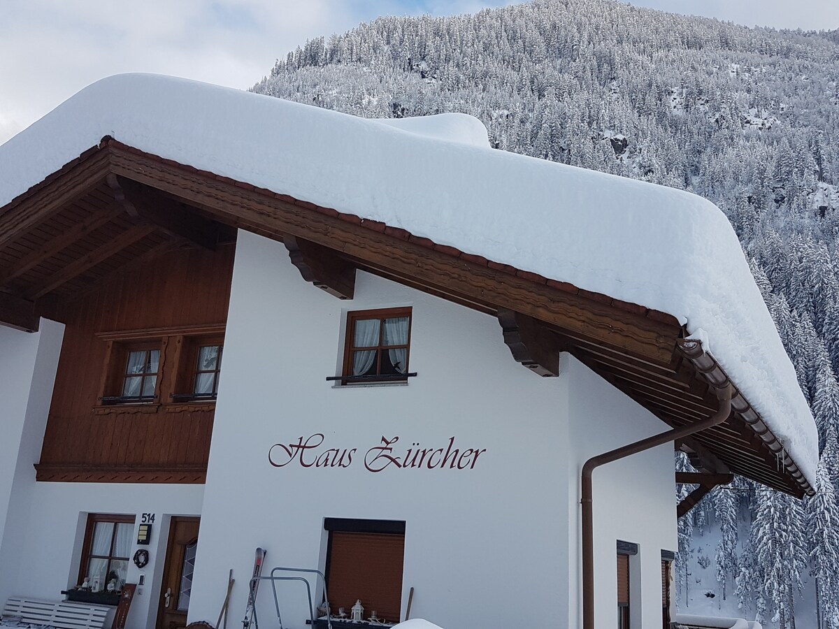 The exterior of Haus Zürcher is shown, featuring a traditional Alpine style with a sloped roof covered in fresh snow. Large windows reflect the serene winter landscape, while a backdrop of snow-covered mountains is visible, enhancing the tranquil atmosphere.