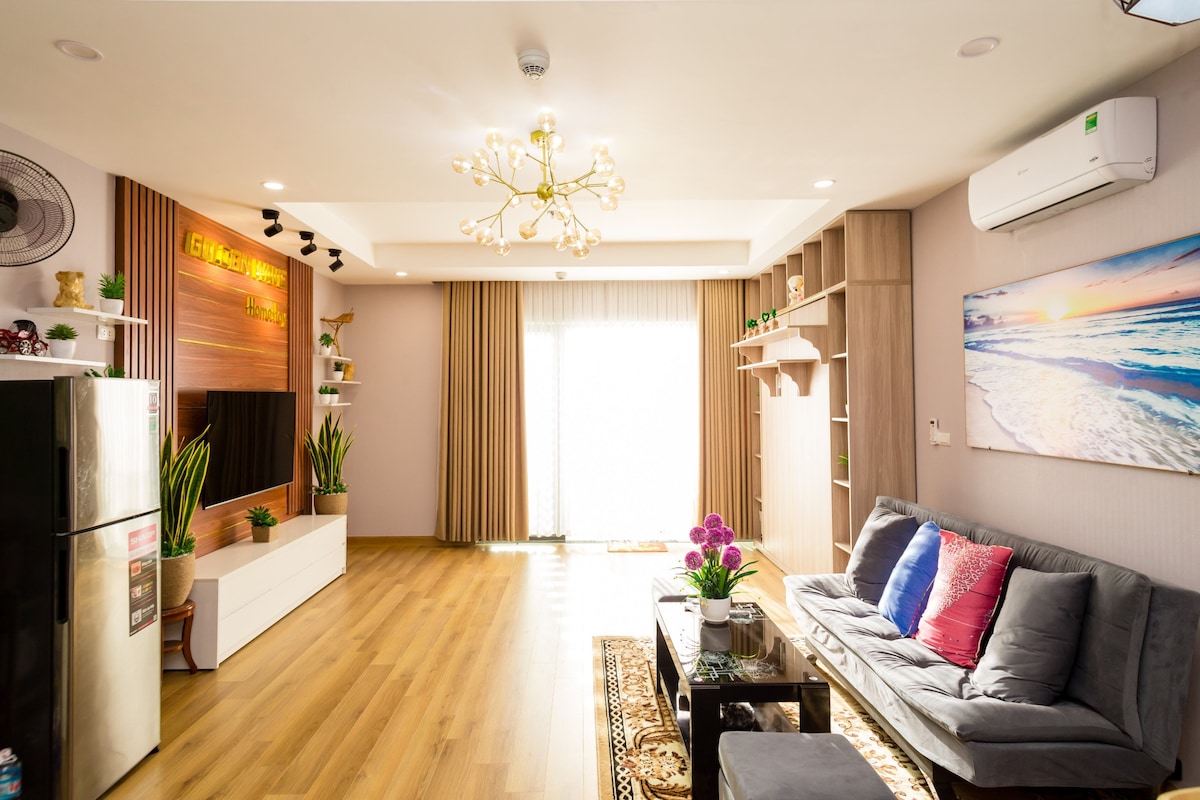 A spacious living area features a comfortable gray sofa adorned with colorful throw pillows. A sleek coffee table sits in front, while plants and decorative shelving add a fresh touch. Large windows allow natural light to fill the room, and air conditioning ensures comfort.