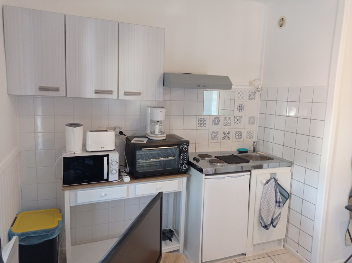 The kitchen features essential appliances including a microwave, coffee maker, toaster oven, and refrigerator. Storage cabinets are present above the countertop, which is complemented by a sink and a stove. White tiles cover the walls, adding a clean and functional appearance.