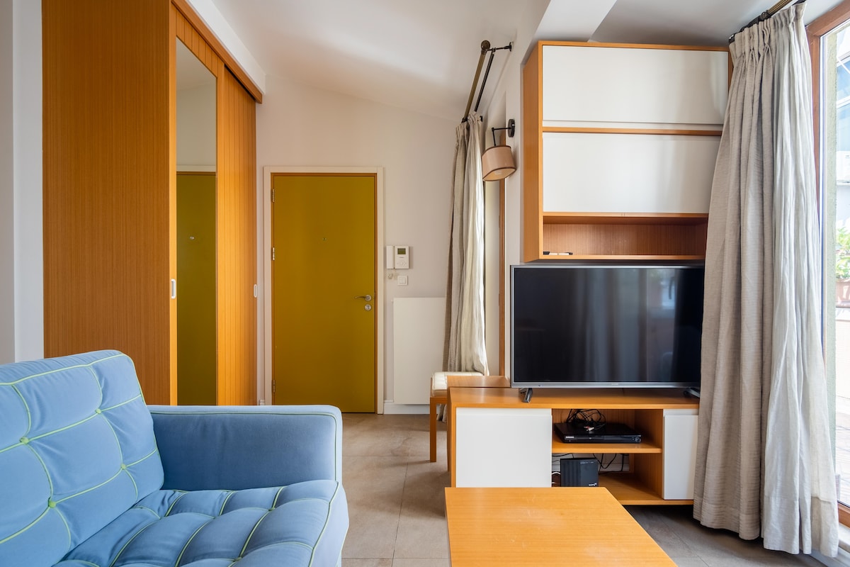 A living area includes a blue couch and a wooden coffee table, with a flat screen TV positioned on a light wood console. Natural light filters through sheer curtains, and an orange door adds a pop of color to the scene.