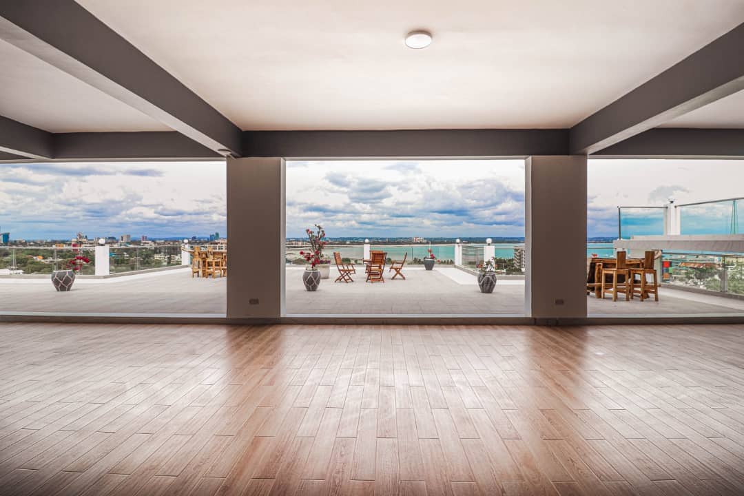 A spacious area features large glass doors that open to an expansive outdoor terrace. The terrace is furnished with wooden tables and chairs, providing views of the ocean and city skyline. Natural light fills the room, highlighting the warm wooden floor.