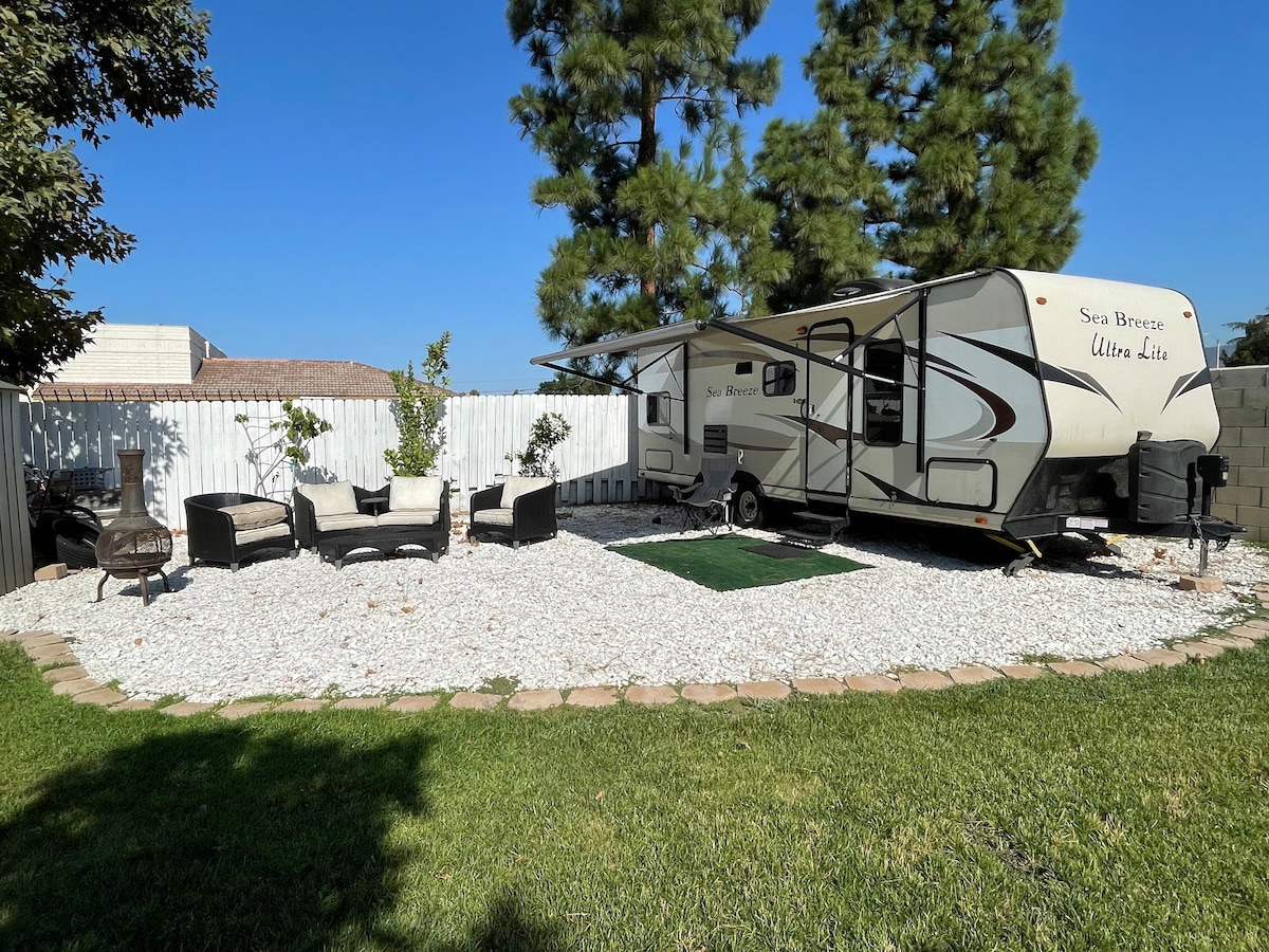A spacious RV is parked on a gravel surface within a landscaped area featuring green grass. A seating arrangement with comfortable chairs and a fire pit is positioned under a shaded awning, providing a relaxing outdoor space surrounded by low-maintenance plants.