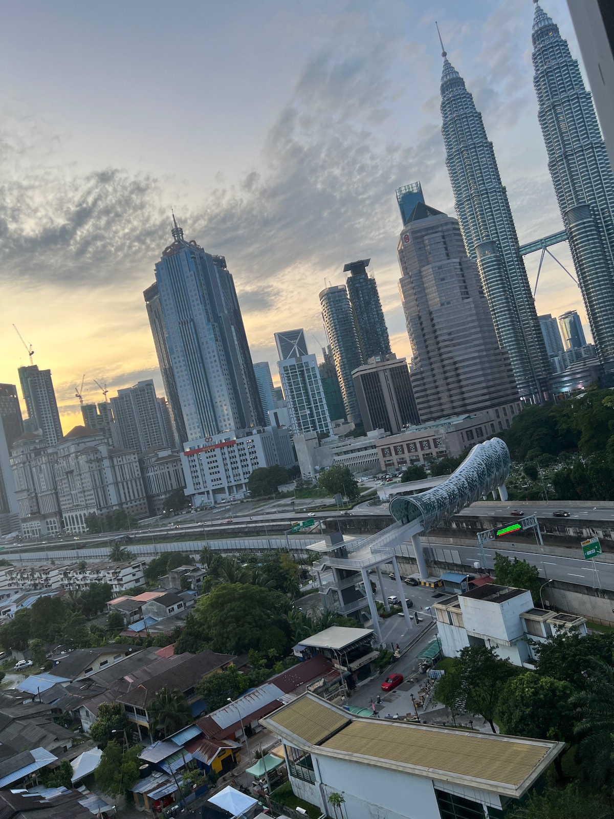The vibrant cityscape features the iconic Petronas Towers, surrounded by a mix of modern skyscrapers and traditional houses. A unique bridge is visible connecting the area, with greenery and roads below. The sky exhibits a blend of soft orange and blue hues during sunset.