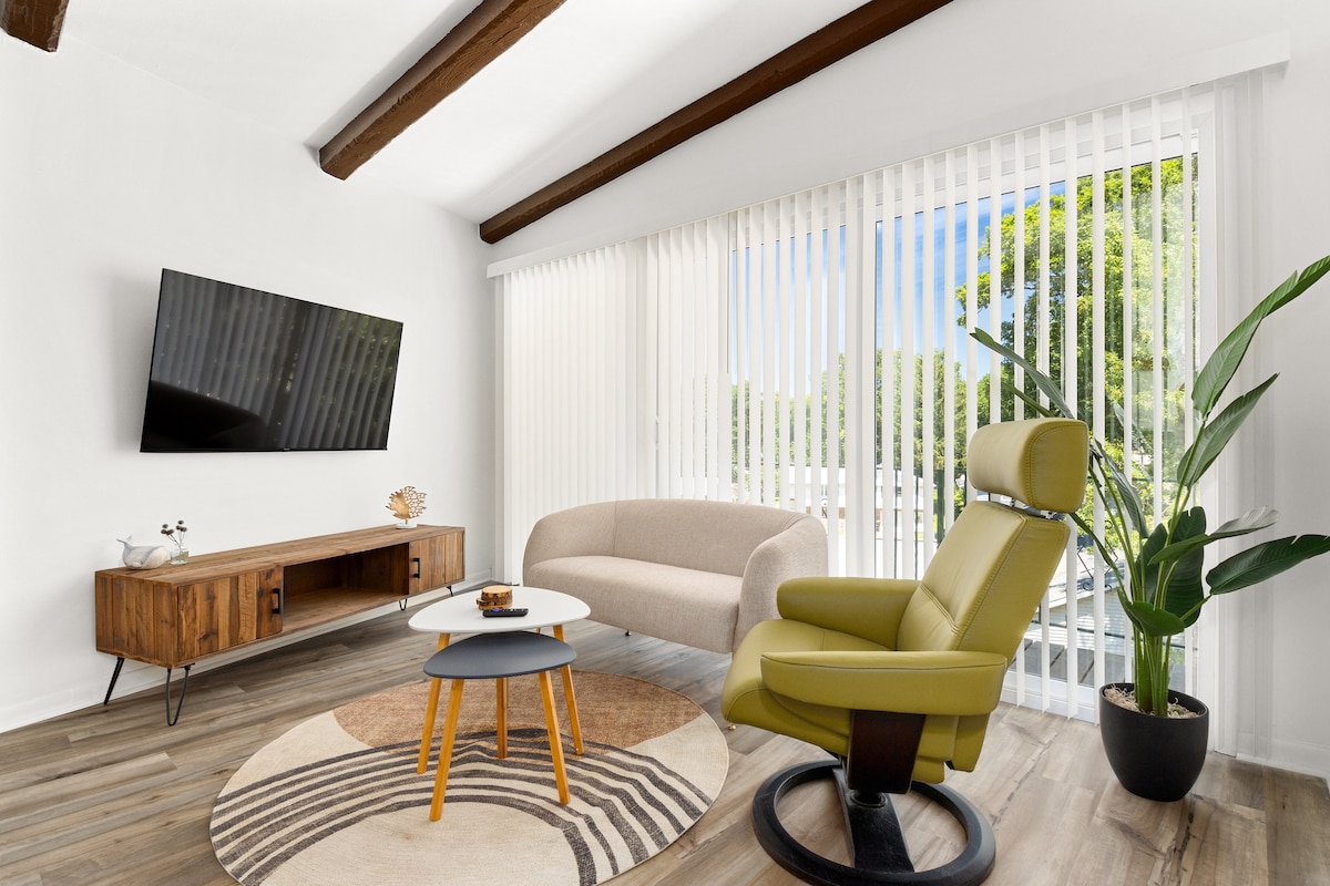 The living area features a comfortable beige sofa paired with a light green armchair, positioned beside a round coffee table on a patterned rug. A large wall-mounted television is present, with natural light filtering through vertical blinds and a potted plant adds a touch of greenery.