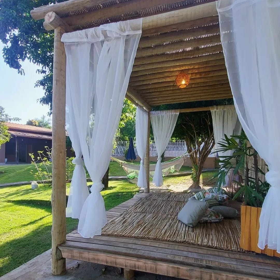 A cozy outdoor seating area is framed by light, flowing curtains. A natural wood structure provides shelter, while a hammock is visible for relaxation. Lush greenery surrounds the space, adding a touch of nature and tranquility, with comfortable cushions arranged on the wooden platform.