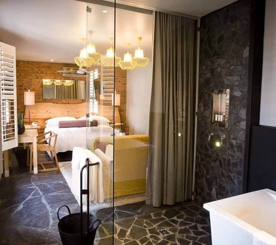 A modern bathroom features a glass partition overlooking a spacious bedroom. Natural stone walls and dark flooring create an elegant contrast. A freestanding bathtub is positioned near the shower, while soft lighting from pendant fixtures illuminates the area, enhancing the cozy ambiance.