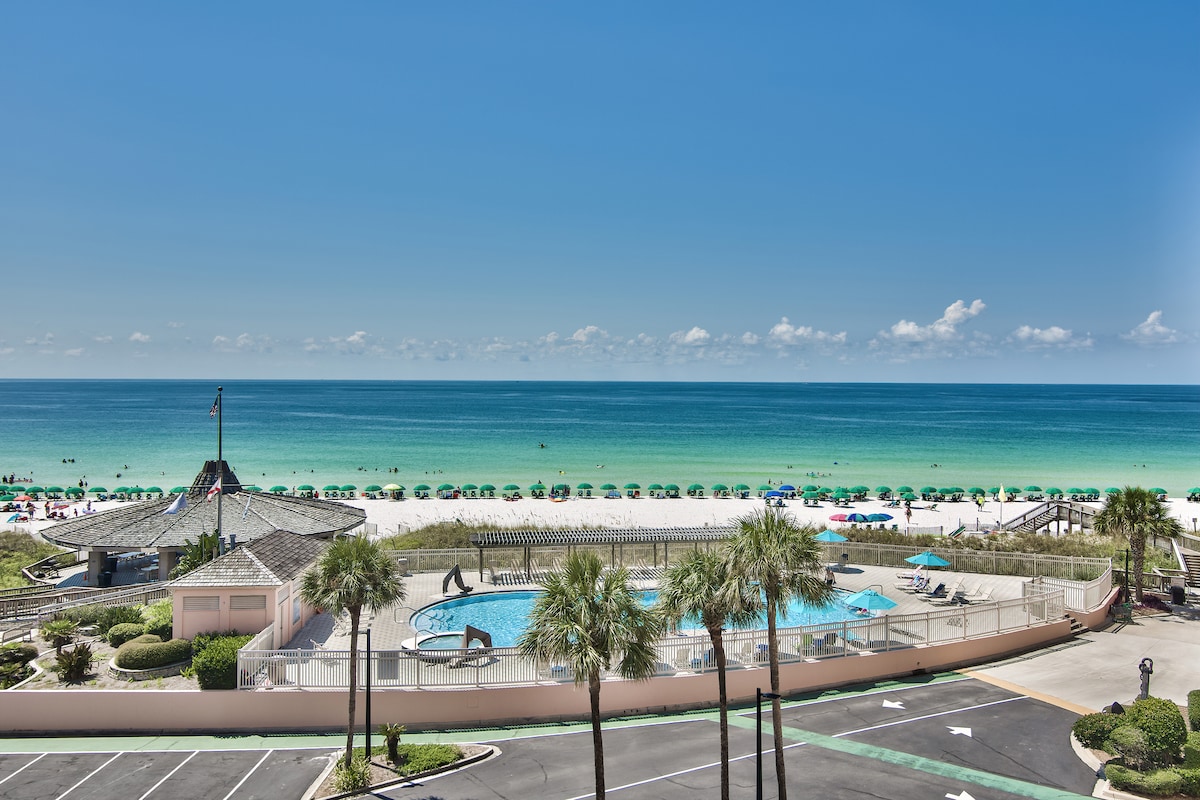 A view showcasing the vibrant emerald waters of the Gulf and a sandy beach lined with sun umbrellas. A swimming pool is visible, surrounded by lounge chairs, offering a relaxing space. Clear blue skies extend above, completing the coastal landscape.