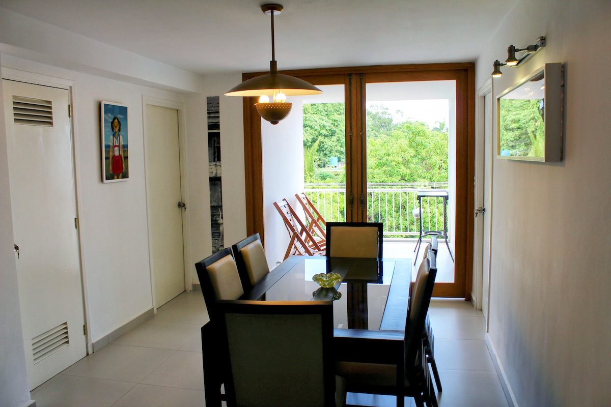 A dining area is presented with a rectangular table surrounded by six upholstered chairs. Large glass windows allow natural light to illuminate the space, with a view of greenery visible outside. Decorative wall art adds a touch of color to the bright, neutral-toned interior.