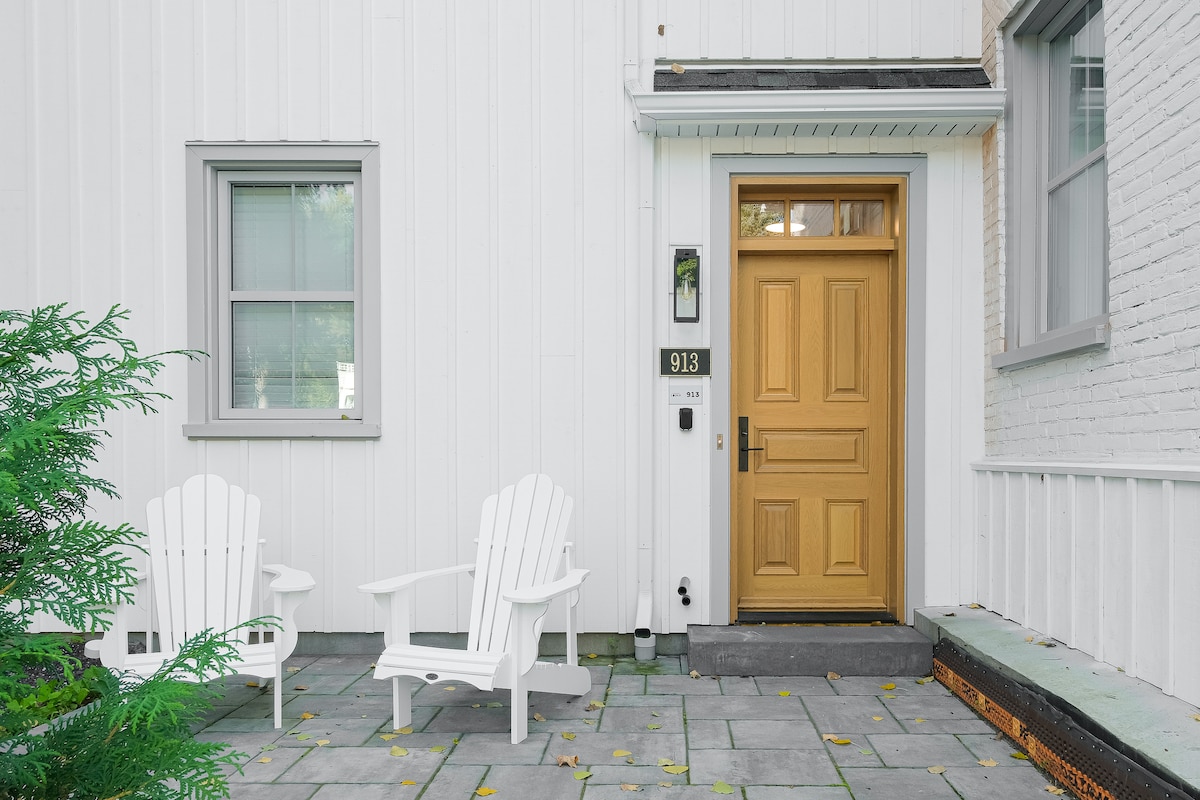 A welcoming entrance features a wooden door with a clear glass panel, flanked by two white Adirondack chairs on a stone patio. Green foliage partially borders the space, enhancing the inviting atmosphere of the entryway.