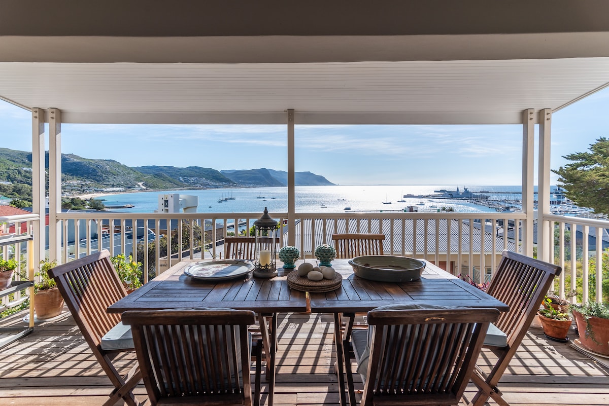 A spacious outdoor dining area is displayed on a covered balcony, featuring a wooden table surrounded by several chairs. The table is adorned with decorative items, and the backdrop reveals panoramic ocean views, with the coastline and boats visible in the distance.