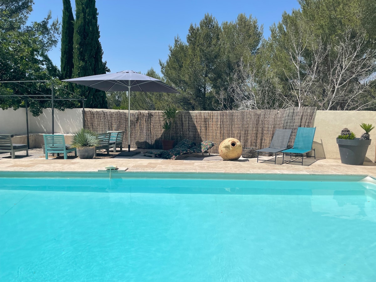 A shared swimming pool is surrounded by a spacious patio area featuring sun loungers and a large umbrella for shade. Potted plants add greenery, and a textured privacy screen enhances the tranquil atmosphere. The clear water reflects the bright blue sky.