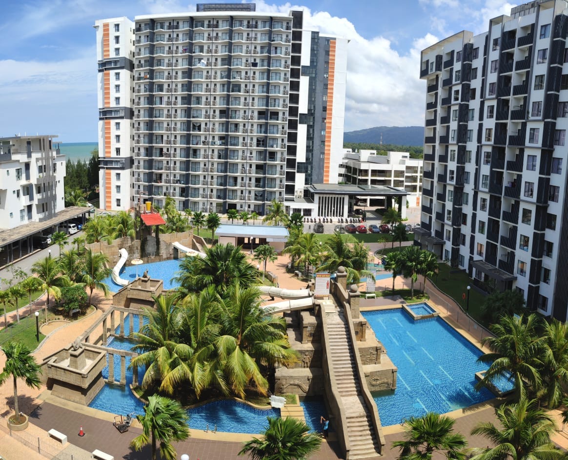 A scenic view of a shared pool area, featuring water slides and lush palm trees. The landscape includes a large swimming pool surrounded by landscaped pathways, with modern apartment buildings in the background, and an expansive sky above.