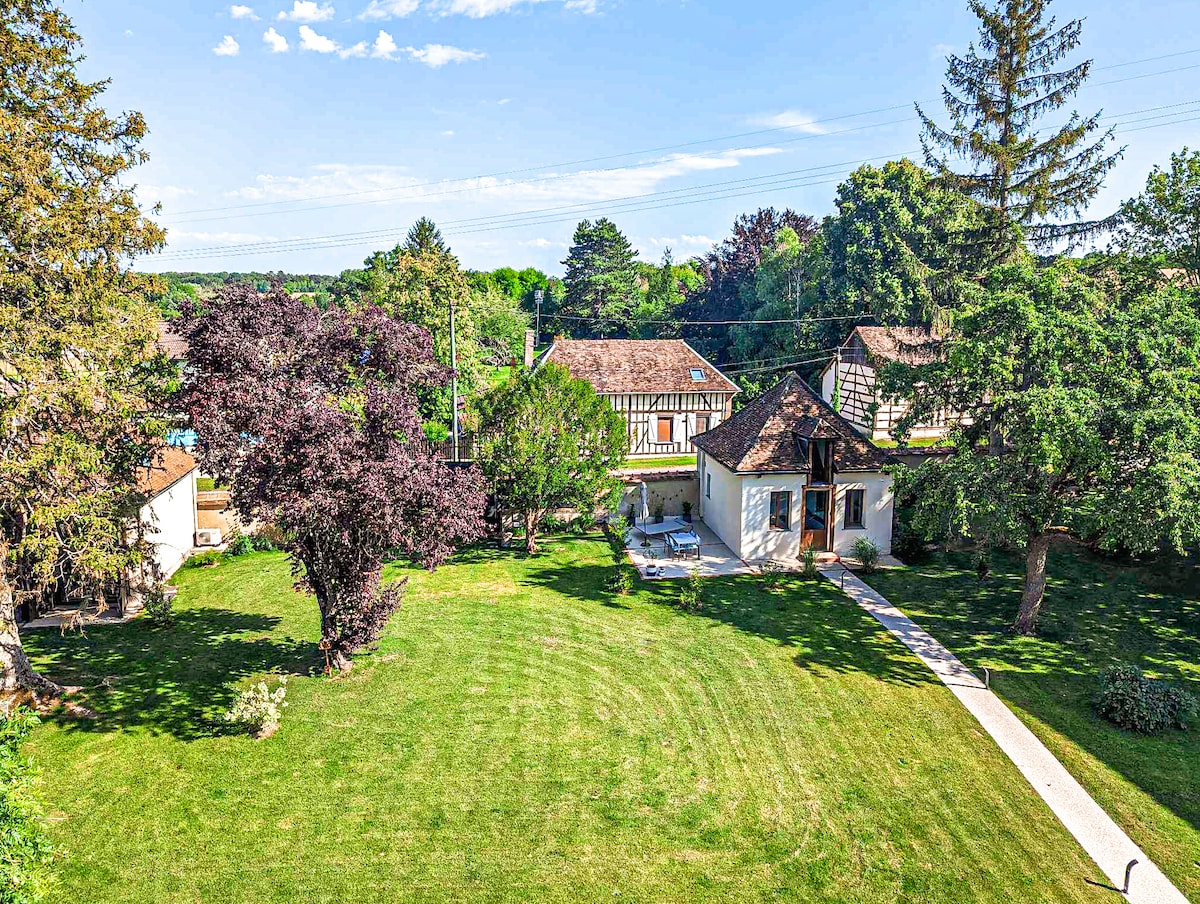 A spacious outdoor area is shown, featuring a well-maintained lawn surrounded by a variety of trees. The view includes several rustic buildings and a pathway leading towards a terrace area, equipped with outdoor seating under the shade of greenery.