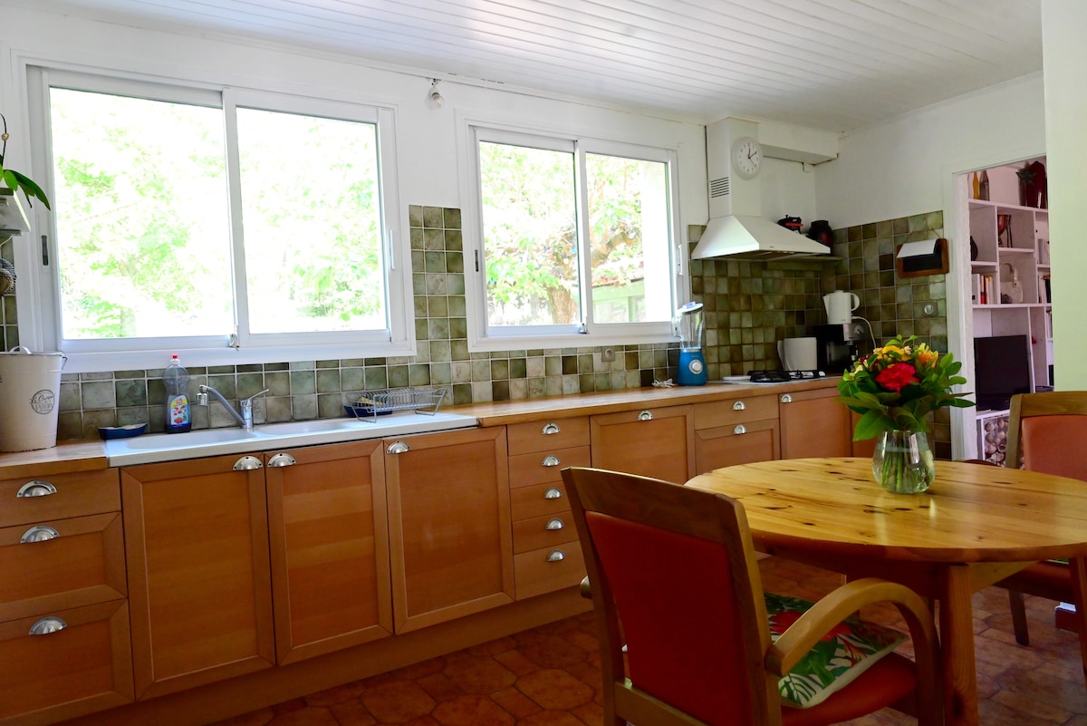 The kitchen is equipped with wooden cabinets and a round dining table surrounded by chairs. Large windows allow natural light to flood the space, offering views of the greenery outside. A stove and various appliances are visible along the counter.