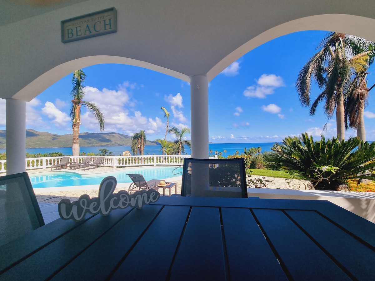A welcoming outdoor seating area is presented under an arched portico. The view reveals a pool surrounded by palm trees and lush greenery, with coastal waters and distant hills visible in the background. Gentle clouds drift across a bright blue sky.