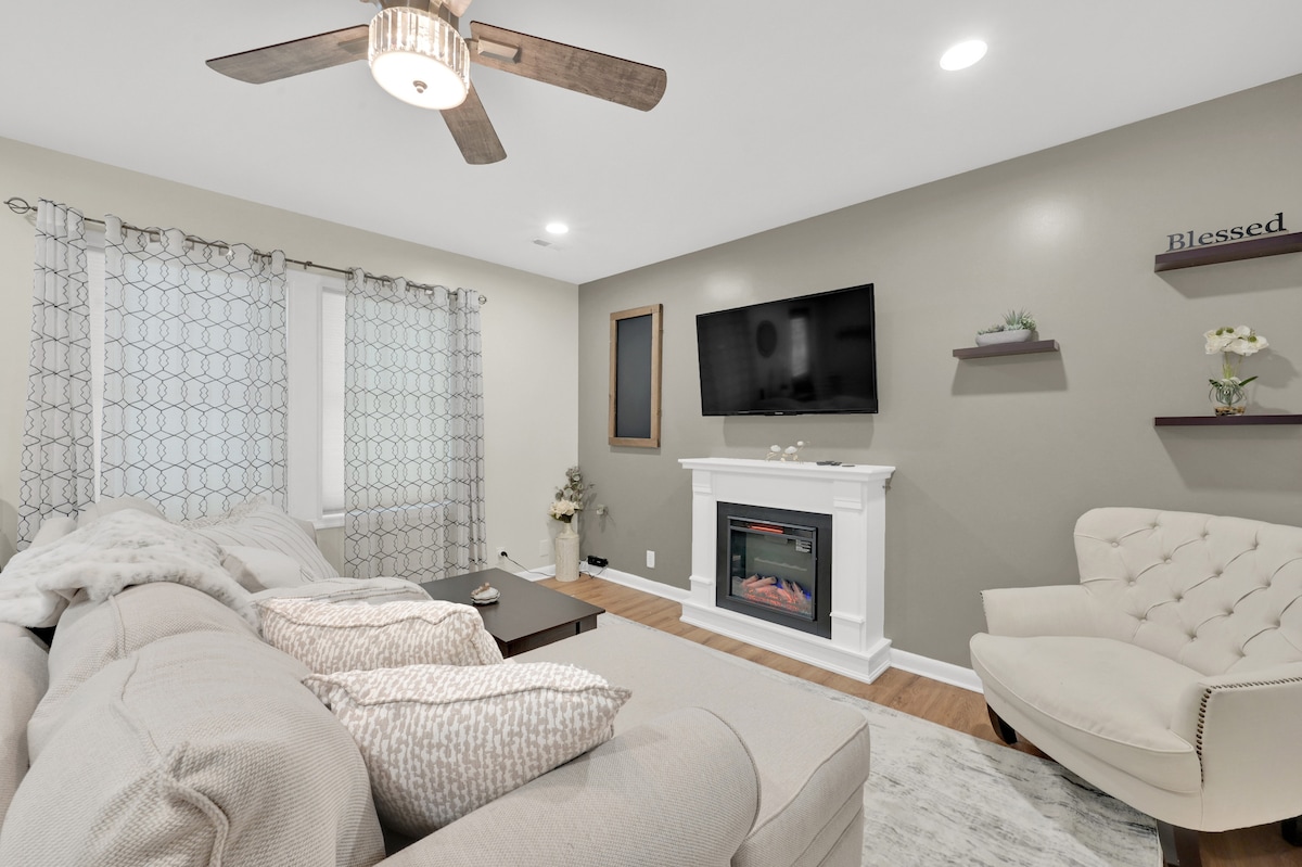 A comfortable living room showcases a light-colored sectional sofa adorned with decorative pillows. A modern fireplace is centered beneath a wall-mounted television. Natural light filters through sheer curtains framing two windows. A stylish accent chair and minimalist shelves enhance the welcoming atmosphere.