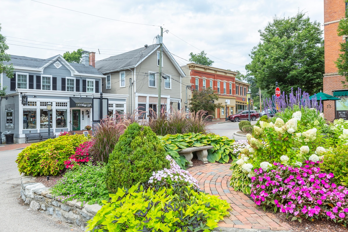 A vibrant garden is showcased, featuring an arrangement of colorful flowers, including pinks and whites, alongside lush green plants. A brick pathway leads to a stone bench, while charming buildings can be seen in the background, contributing to the neighborhood's inviting atmosphere.