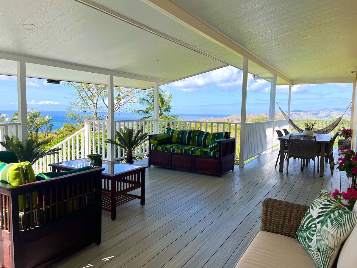 A spacious covered terrace is furnished with comfortable seating, surrounded by vibrant greenery. Tables and chairs are set up for dining, while panoramic ocean views are visible in the distance, inviting relaxation and outdoor enjoyment.