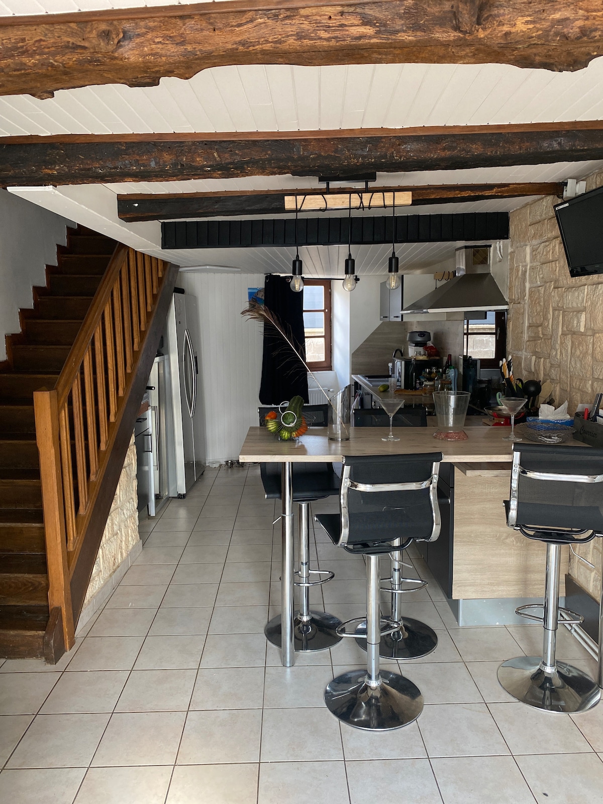 The kitchen area features a modern design with a central island and four black barstools. Visible are stone walls and wooden beams that add character. Appliances, including a refrigerator and stovetop, are positioned thoughtfully. Natural light filters in through a nearby window, enhancing the open space.