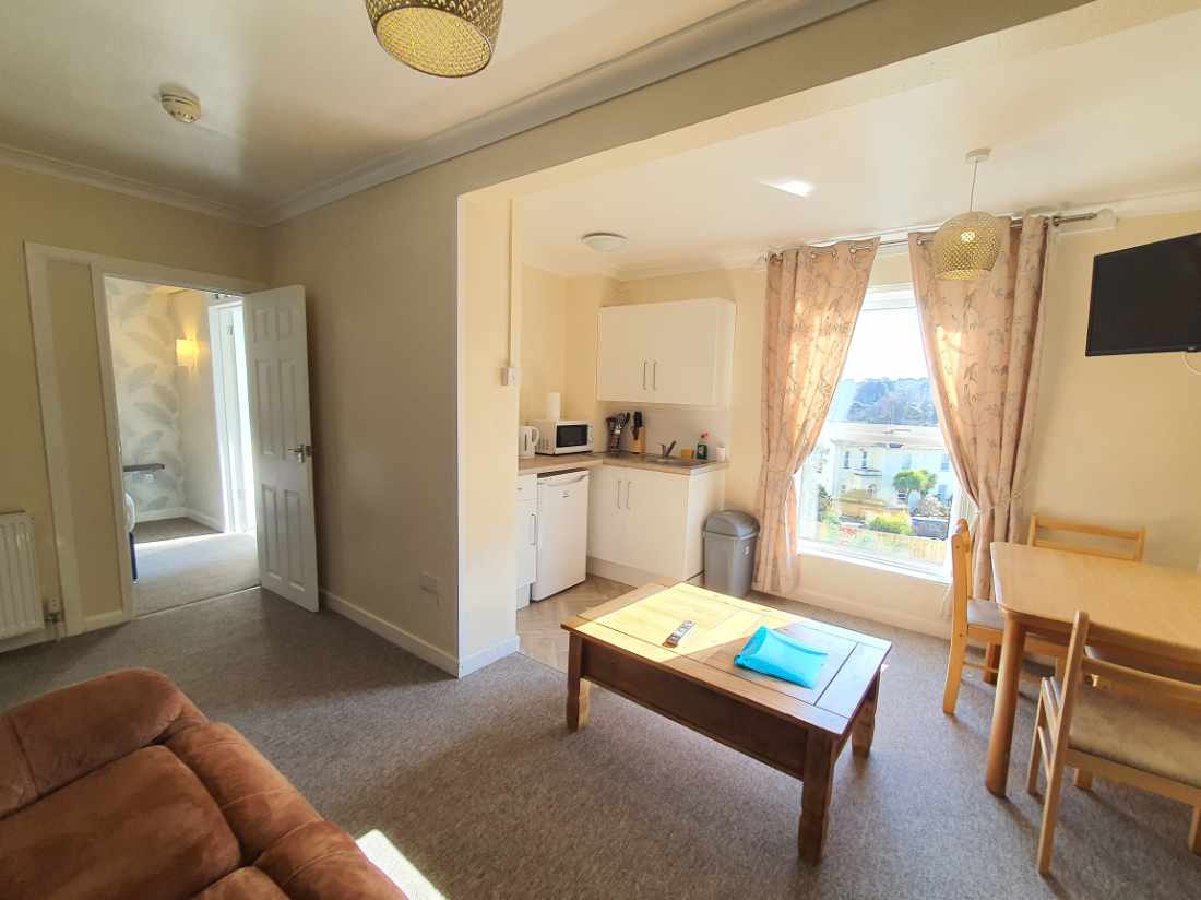 A well-lit living area features a comfortable couch, a wooden coffee table, and a dining set. Natural light enters through a large window adorned with soft curtains, highlighting the adjacent kitchenette, which is equipped with basic appliances.