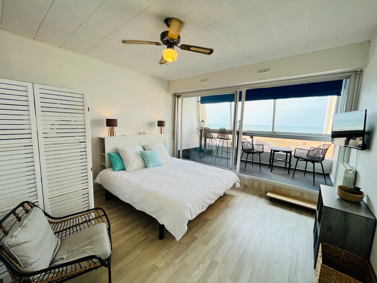 A spacious sleeping area features a large bed dressed in white linens. Natural light filters through large glass doors leading to a closed balcony with ocean views. Two stylish chairs and lamps are positioned at the foot of the bed, while a wall-mounted flat-screen TV is visible.