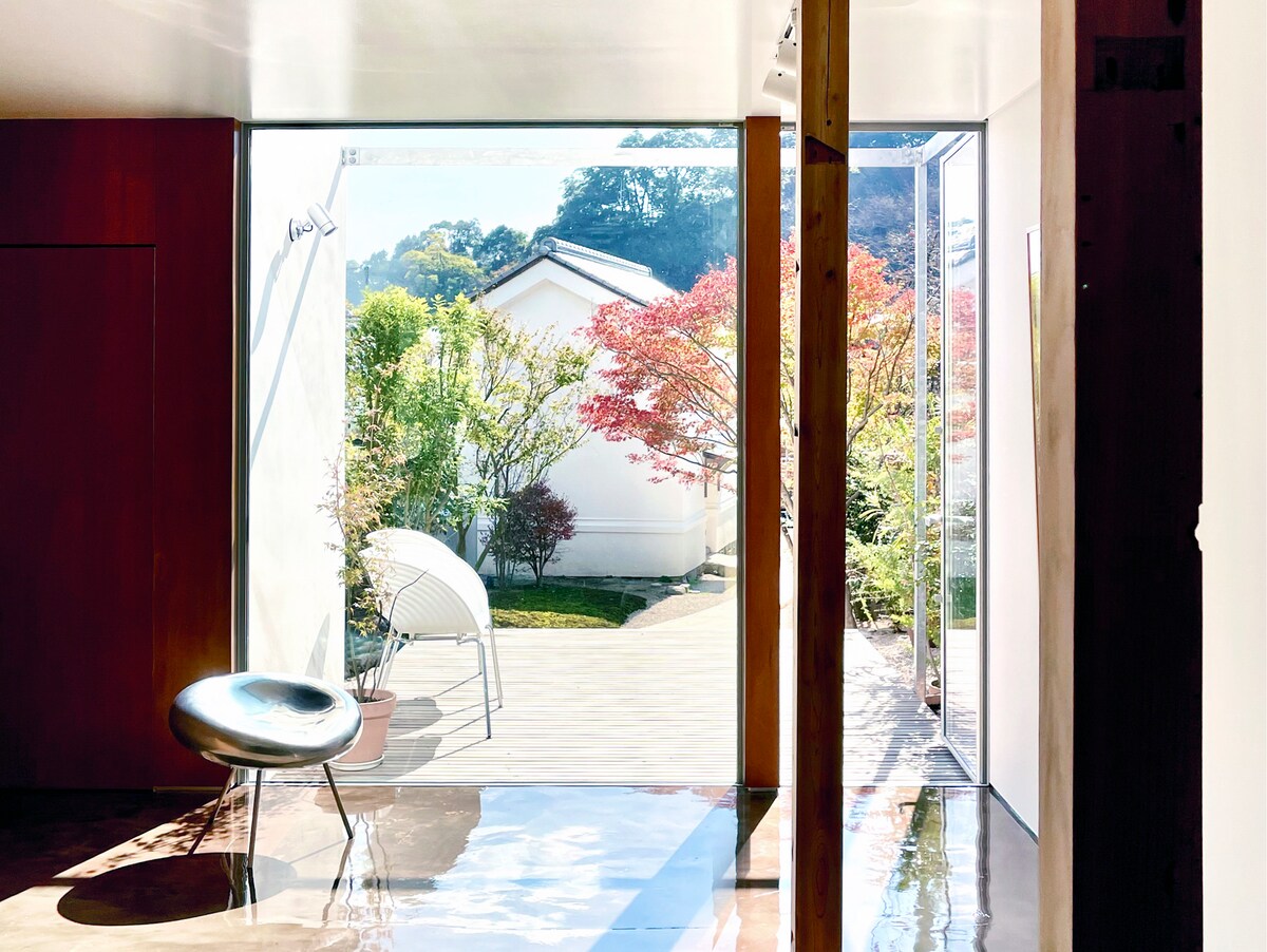 Natural light filters through expansive glass doors, illuminating the interior space. A sleek chair is positioned beside the entrance, offering a view of the garden's vibrant foliage and wooden deck beyond. The connection between indoors and outdoors is enhanced by the surrounding greenery.