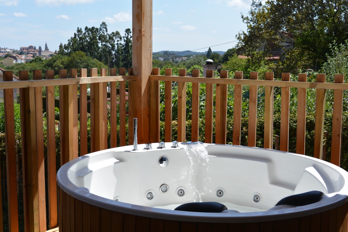 A private terrace features a circular jacuzzi, with water cascading from its spout. Surrounding greenery and a wooden fence provide privacy, while distant views of the city and hills are visible in the background under a clear blue sky.