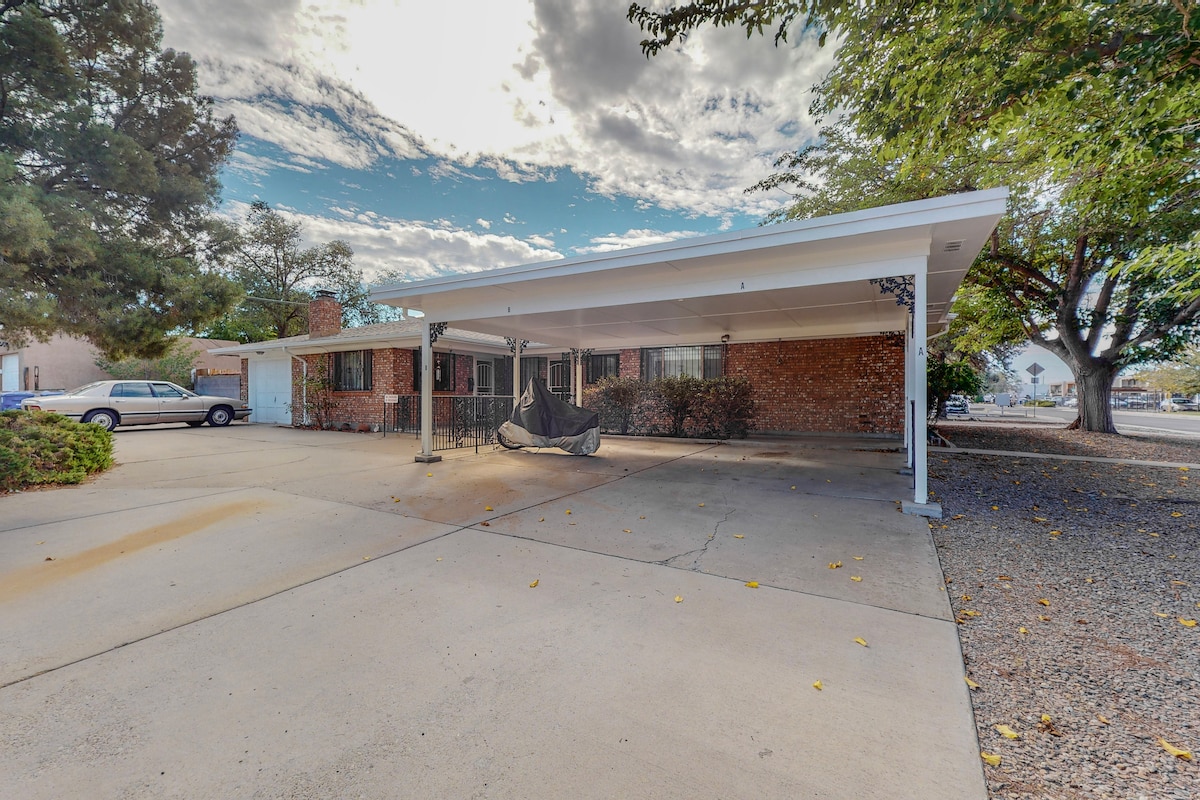 A covered parking area is featured in front of a brick house, providing protection from the elements. The flat surface is spacious and free of obstructions. Surrounding trees offer natural shade, and a gravel area is visible to the side.