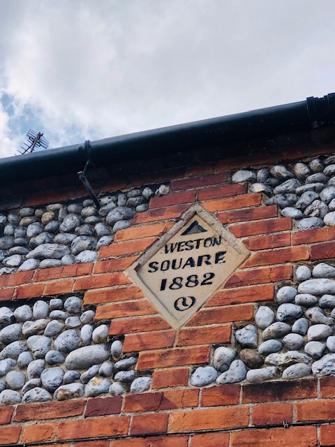 A close-up view of the exterior wall of the cottage, showcasing traditional Norfolk brick and flint construction. A diamond-shaped plaque is embedded in the wall, reading 'WESTON SQUARE 1882,' highlighting the building's historic character.