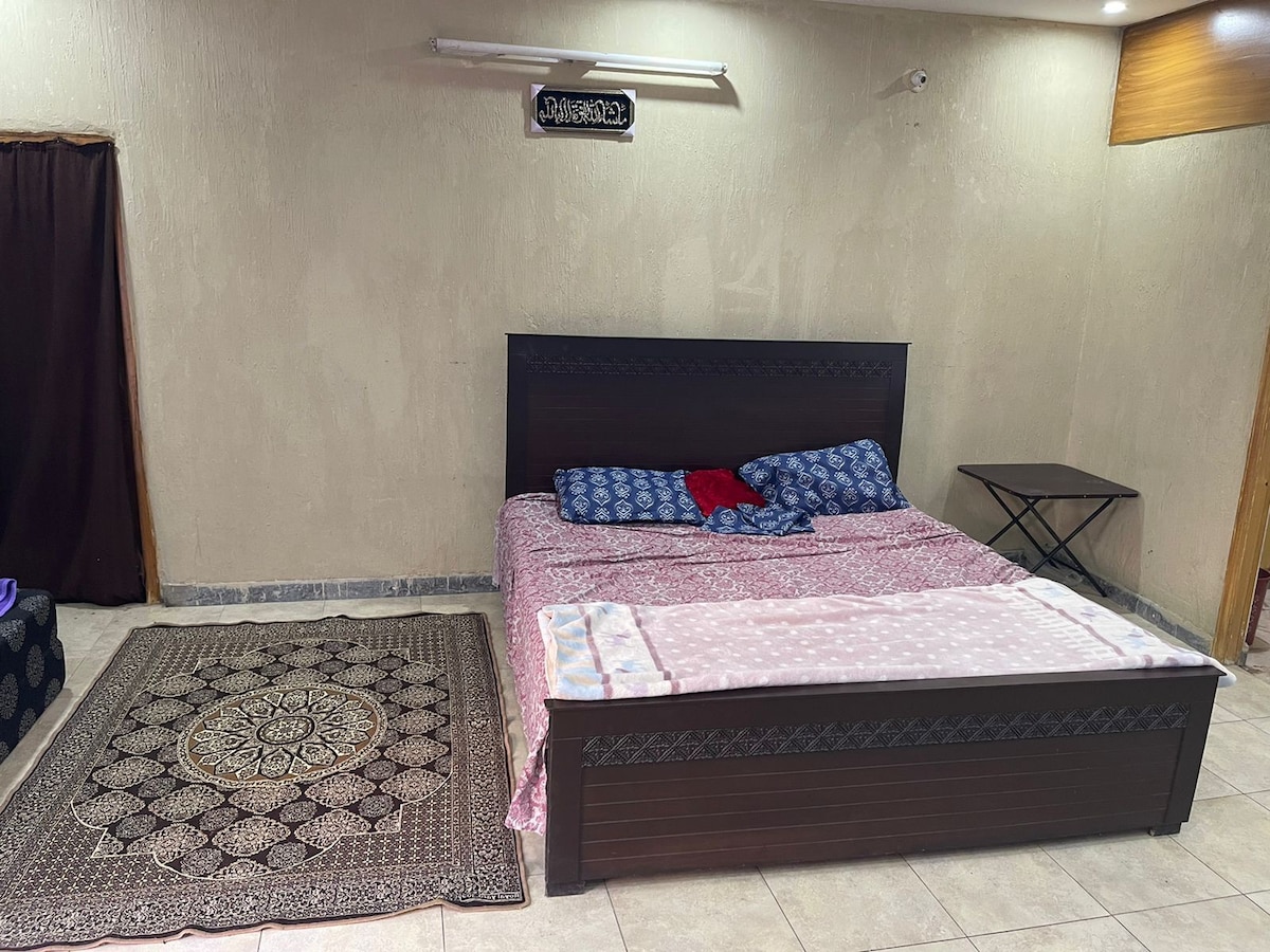 A sleeping area features a dark wooden bed with patterned bedding and two decorative pillows. Beside the bed, a small table is positioned. A large area rug is placed on the tiled floor, complementing the room's neutral walls and soft lighting.