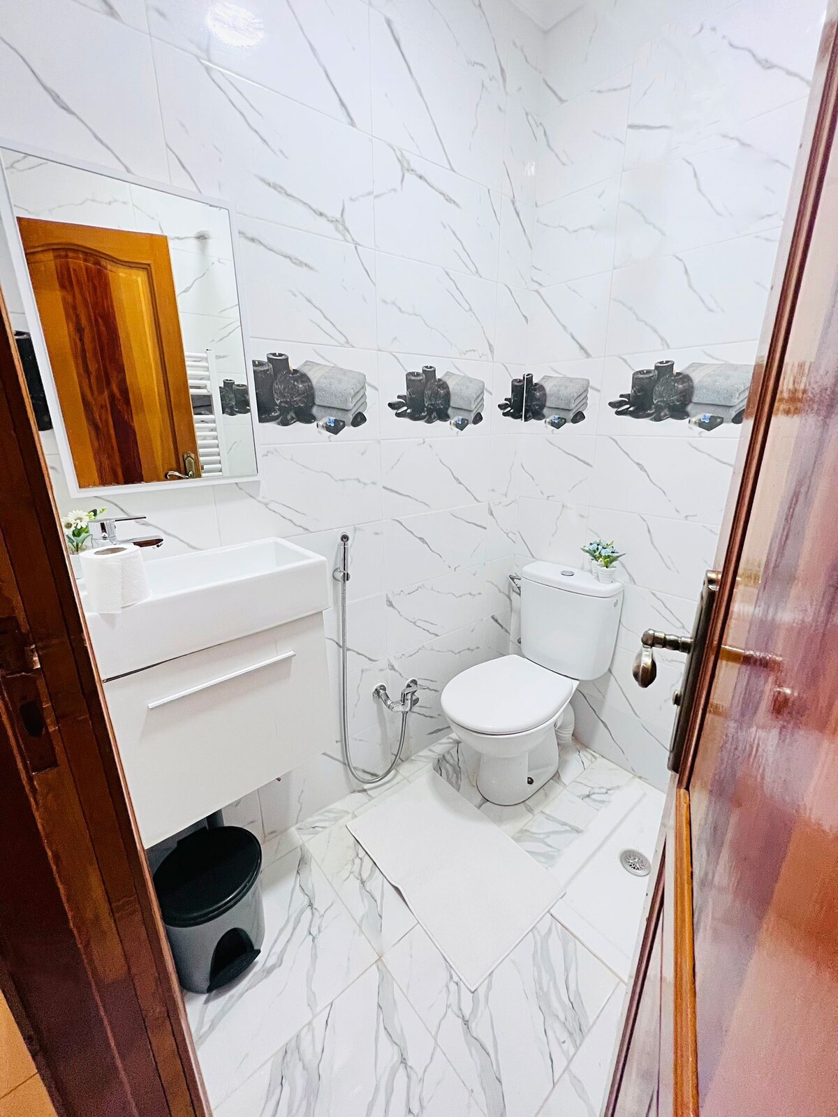 A modern bathroom features white marble walls and a compact design. A sleek vanity with storage is positioned beside a full-length mirror. A toilet and a small waste bin are included, with a soft bath mat on the floor, adding functionality to the space.