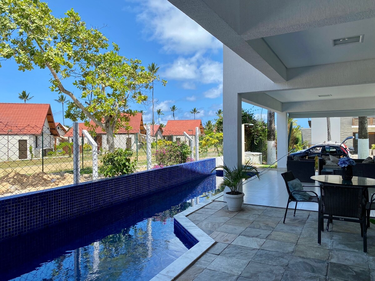 A serene outdoor space features a rectangular pool bordered by stone tiles. A dining area with black wicker chairs is positioned nearby, offering views of palm trees and neighboring structures. The bright sky complements the lush greenery surrounding the area.