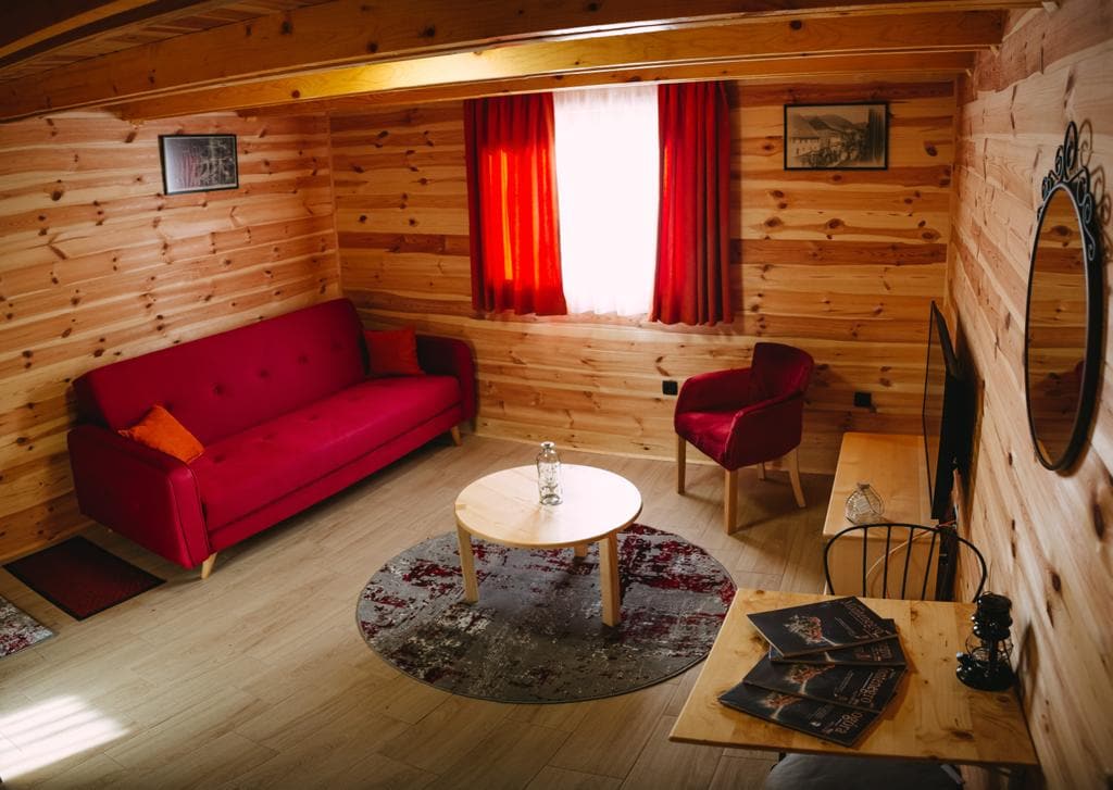 A spacious living area is adorned with warm wooden walls, featuring a red sofa and a coordinating armchair. A circular table sits in the center on a patterned area rug. A large window with red curtains allows natural light to illuminate the space.