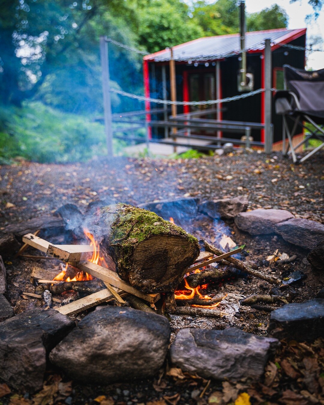 A fire pit surrounded by stones is seen, with flames rising from a log placed atop kindling. In the background, a cabin with a red roof and black walls is partially visible, along with a chair nearby. Soft smoke drifts upward, blending into the natural surroundings.