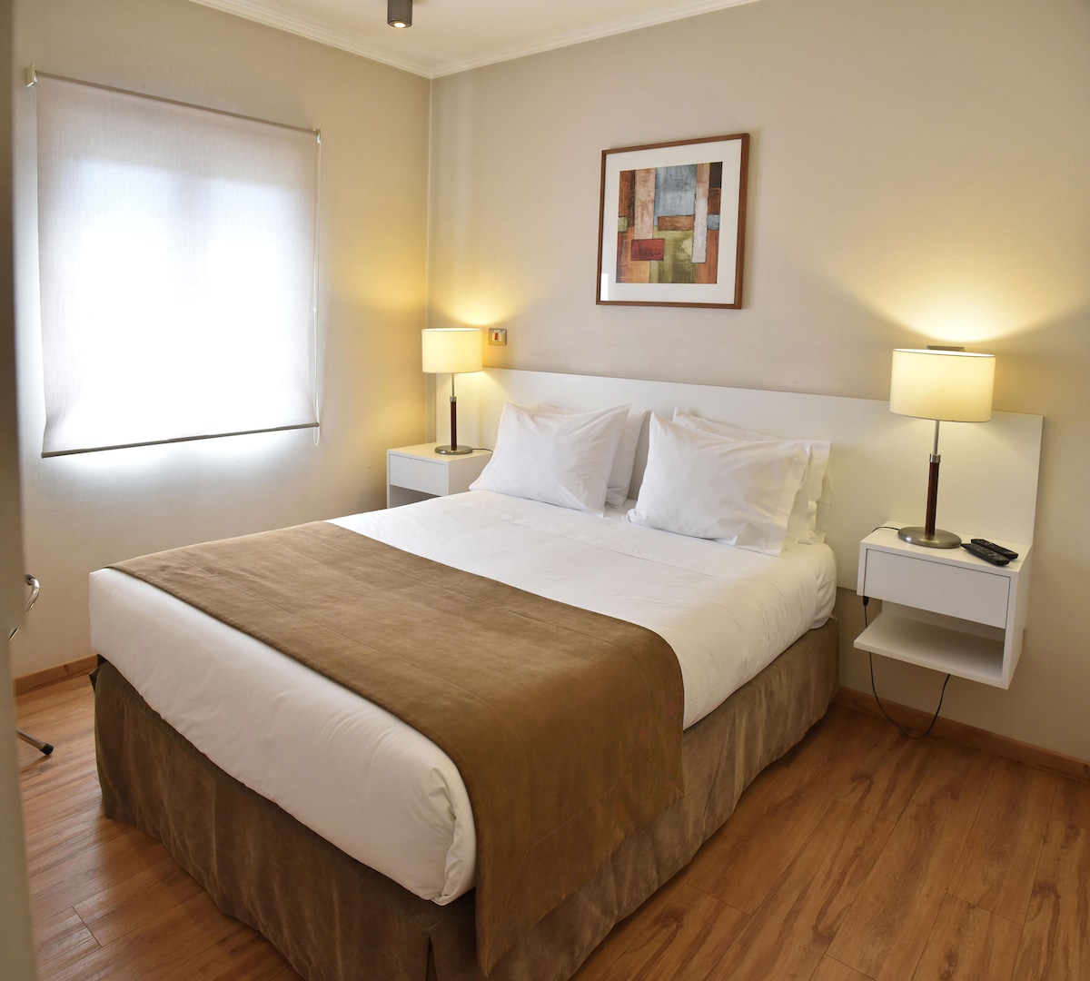 A modern bedroom is presented with a spacious bed featuring white linens. Nightstands with lamps are positioned on either side. Natural light fills the room through a window covered by a light-filtering shade, and a piece of framed wall art adds a subtle touch.