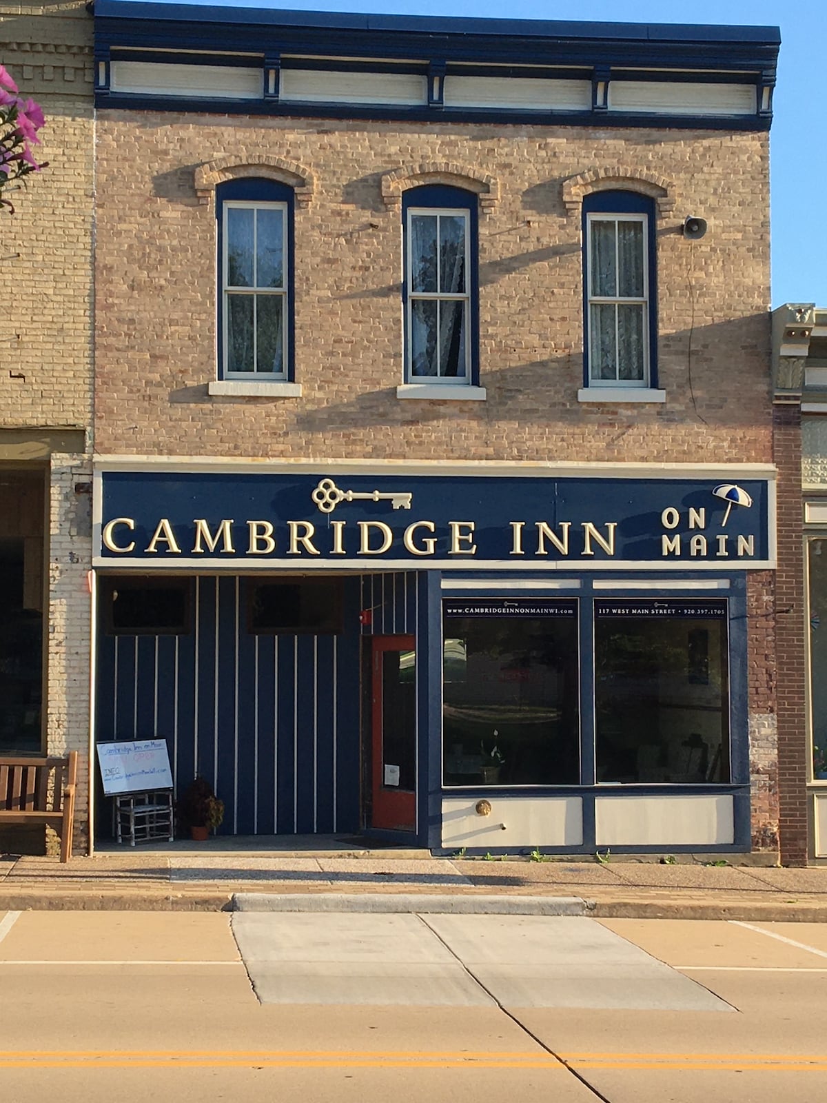 The exterior of the Cambridge Inn on Main features a classic brick facade, adorned with large windows that invite natural light. The blue and white signage is prominently displayed above the entrance, complemented by subtle decorative elements that enhance the building's inviting appearance.