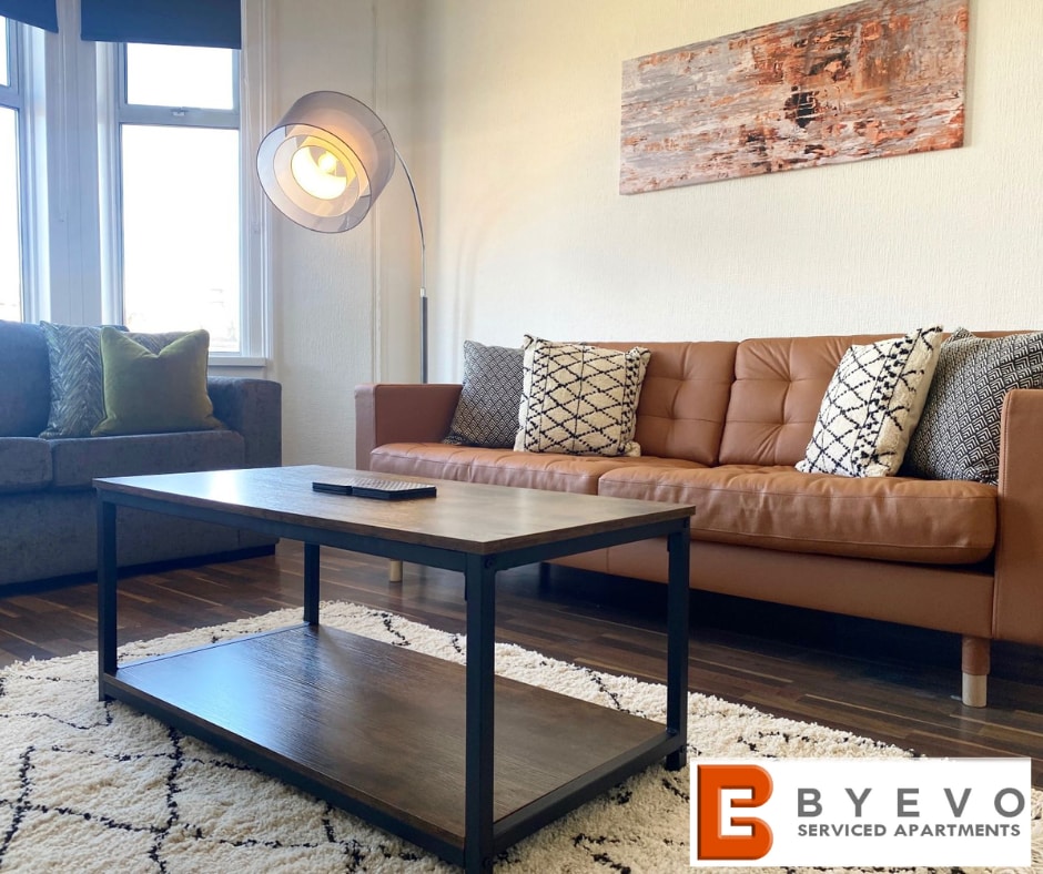A modern lounge area features a stylish brown leather sofa and a plush green couch. A wooden coffee table sits in the center on a soft rug. Natural light streams through large windows, highlighting the contemporary decor and warm tones of the furnishings.