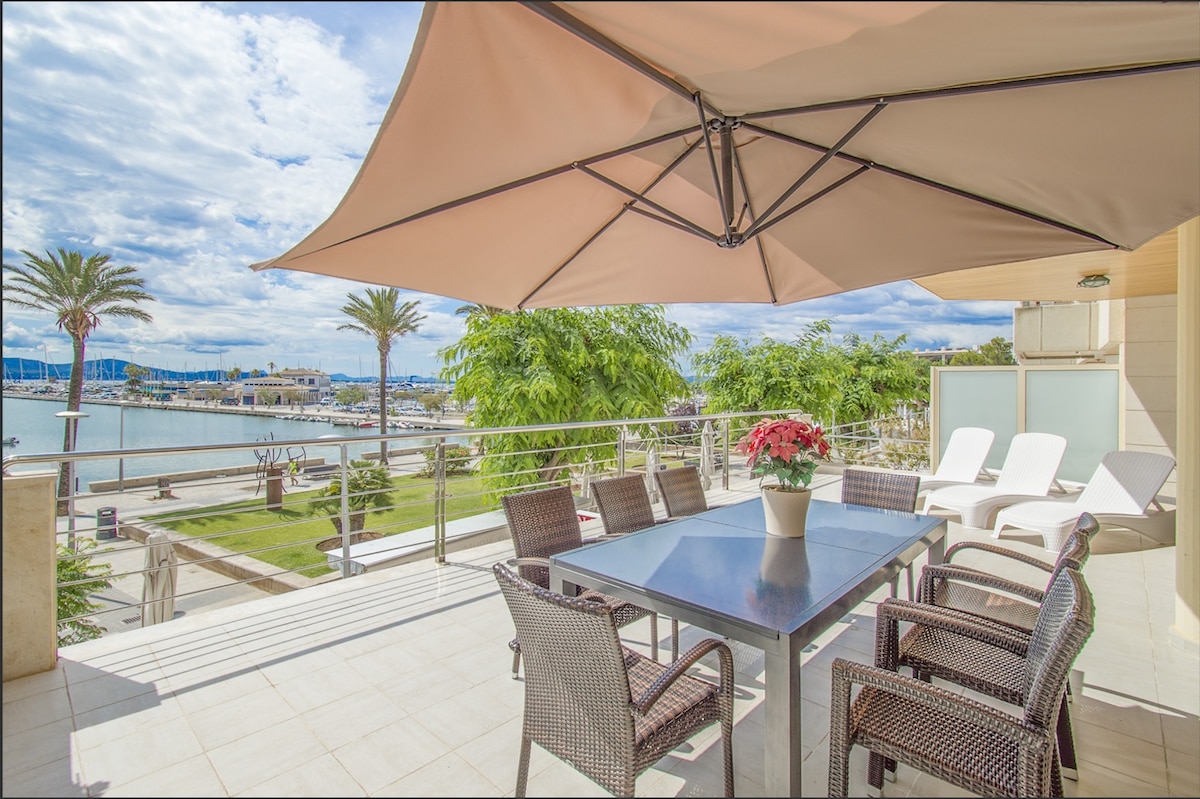 A spacious outdoor terrace features a large table surrounded by several wicker chairs. A sunshade is positioned overhead, providing shade. Potted flowers are present on the table. The terrace offers views of a marina with moored boats and greenery nearby.