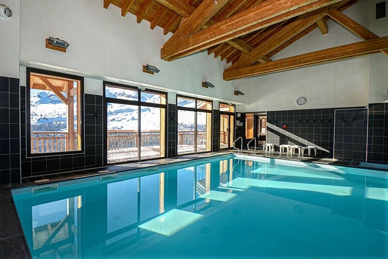A spacious indoor pool area features large windows overlooking the mountains, allowing natural light to fill the space. The pool is surrounded by dark tiles and wooden beams on the ceiling. Several chairs line one side of the pool, providing seating for relaxation.