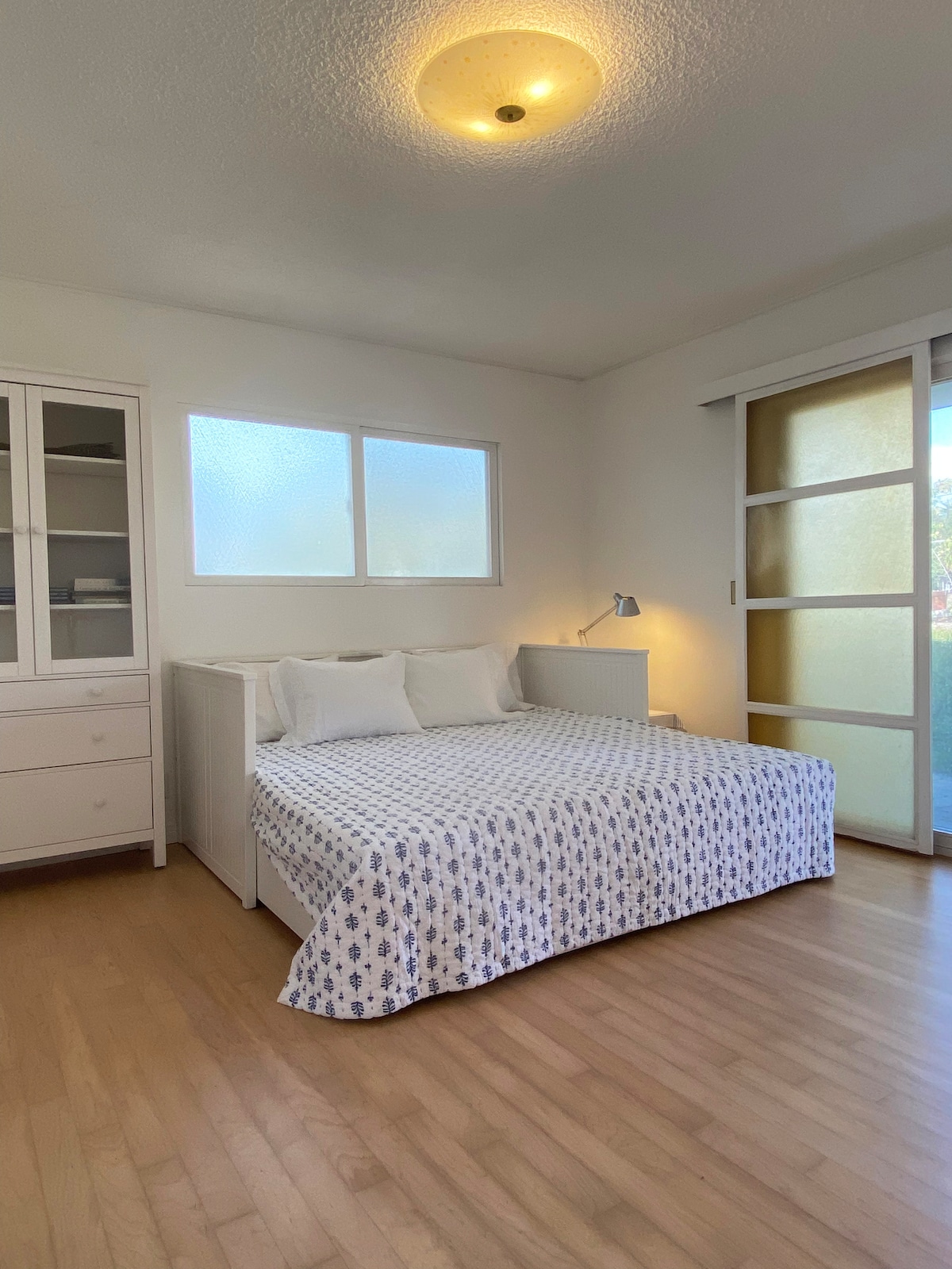 A spacious bedroom features a large bed with a patterned bedspread and multiple pillows. A two-panel sliding door provides access to the outside, while a large window allows natural light to brighten the room. A white cabinet and lamp are positioned beside the bed.