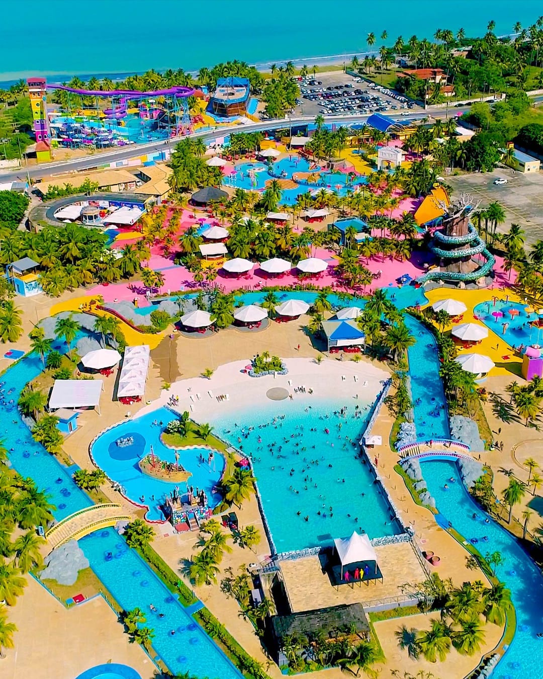 Aerial view of a vibrant water park featuring multiple pools and slides, surrounded by lush palm trees. Colorful structures and shaded cabanas are scattered throughout the area, with people enjoying the various attractions. A portion of the beach and ocean is visible in the background.