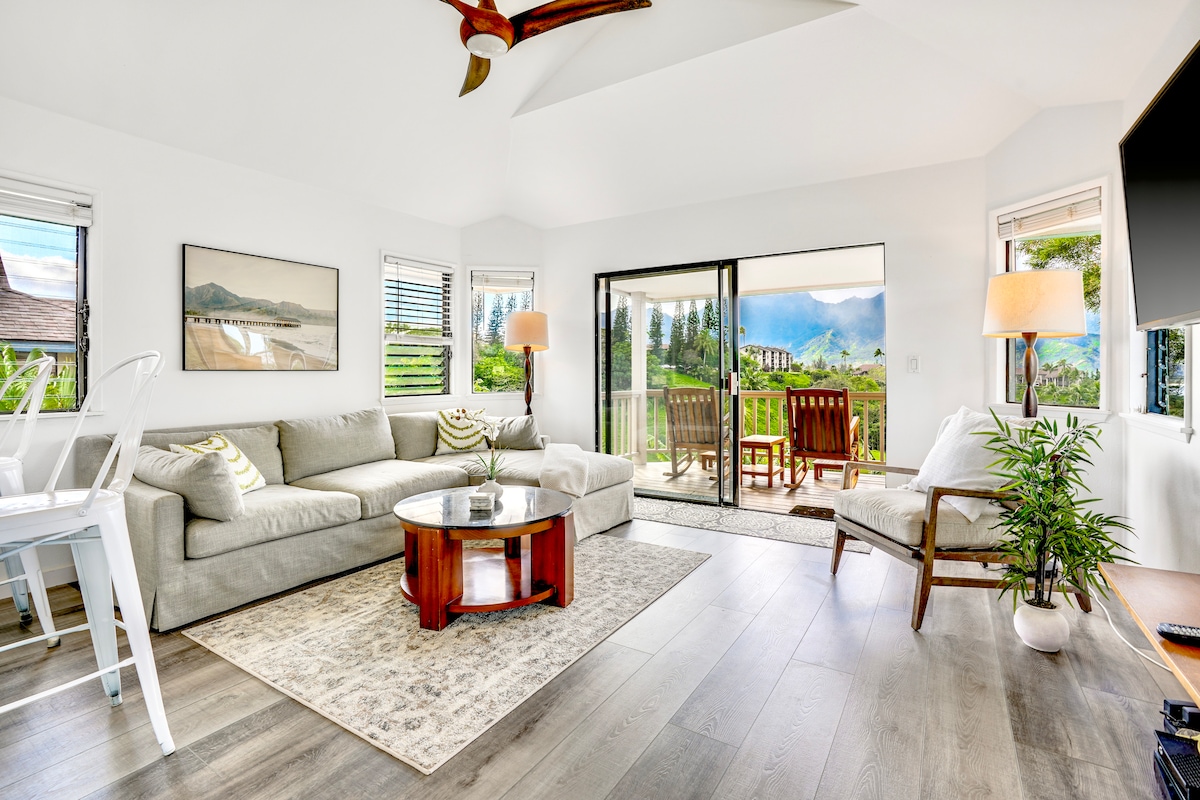 A spacious living room features a large, light-colored sofa and a round wooden coffee table on a soft area rug. Floor-to-ceiling glass doors allow for natural light and views of the outdoors. A wooden dining table is visible on the balcony, alongside a comfortable armchair and floor lamp.